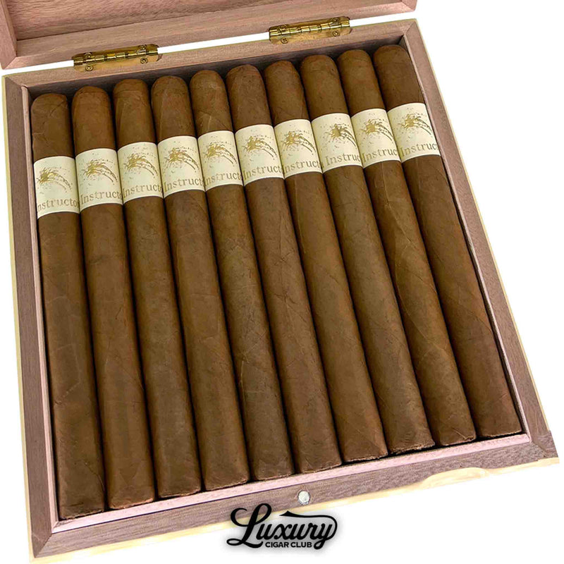 Close-up of La Instructora Delicado Churchill cigars inside a natural wood box showcasing uniform Connecticut shade wrappers and refined cream bands with gold embossed artwork. Luxury Cigar Club logo underneath.