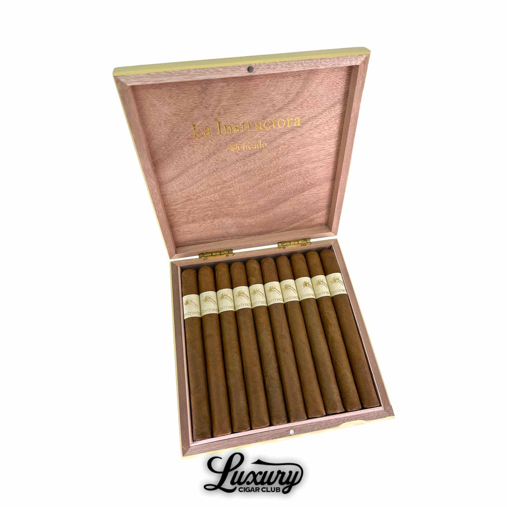 Open wooden box of La Instructora Delicado Churchill cigars displaying neatly arranged cigars with smooth light brown wrappers and elegant cream-colored bands featuring gold detailing. Luxury Cigar Club logo below.
