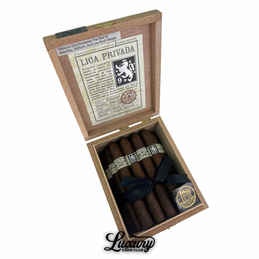 Open box of Liga Privada No. 9 Belicoso Oscuro cigars featuring six dark, tapered cigars with black and white bands, tied with a black ribbon, a Drew Estate coin, and blend details on the inner lid, with Luxury Cigar Club logo below.