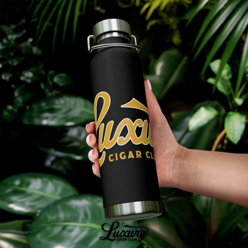 outdoor hand shot of luxury cigar club copper vacuum 22oz insulated bottle black with gold logo surrounded by greenery