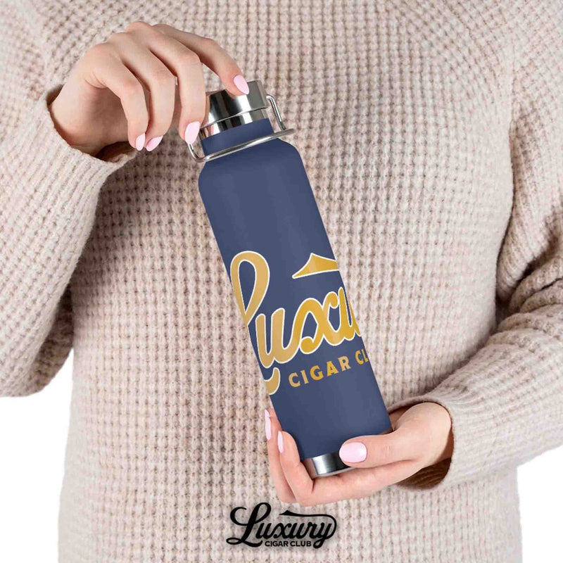Person holding Luxury Cigar Club navy copper vacuum insulated bottle with gold logo while wearing beige sweater