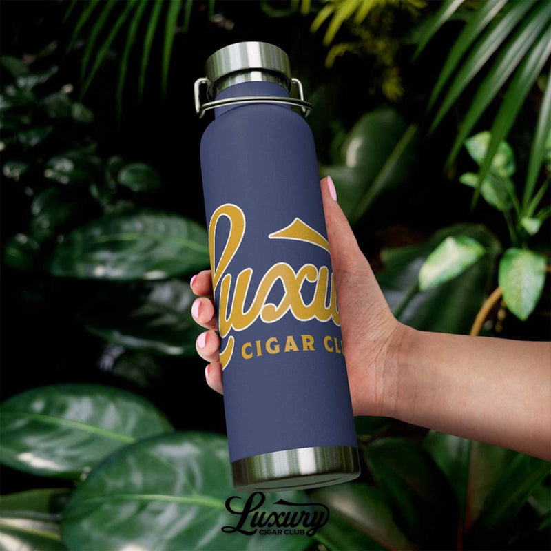 Hand holding Luxury Cigar Club navy insulated stainless steel bottle with gold lettering in outdoor tropical setting with green leaves