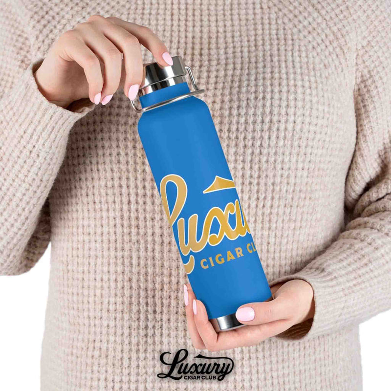 Lifestyle image of person holding Luxury Cigar Club pebble blue insulated water bottle in cozy beige sweater showing gold logo prominently