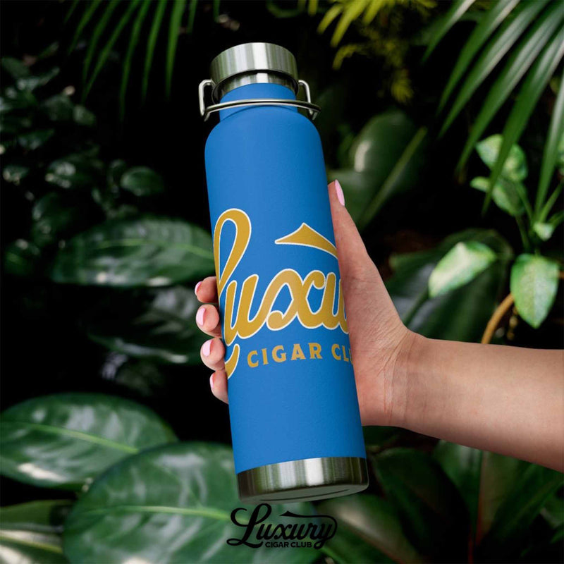 Outdoor lifestyle shot of Luxury Cigar Club pebble blue insulated bottle with gold lettering being held against lush green tropical leaves