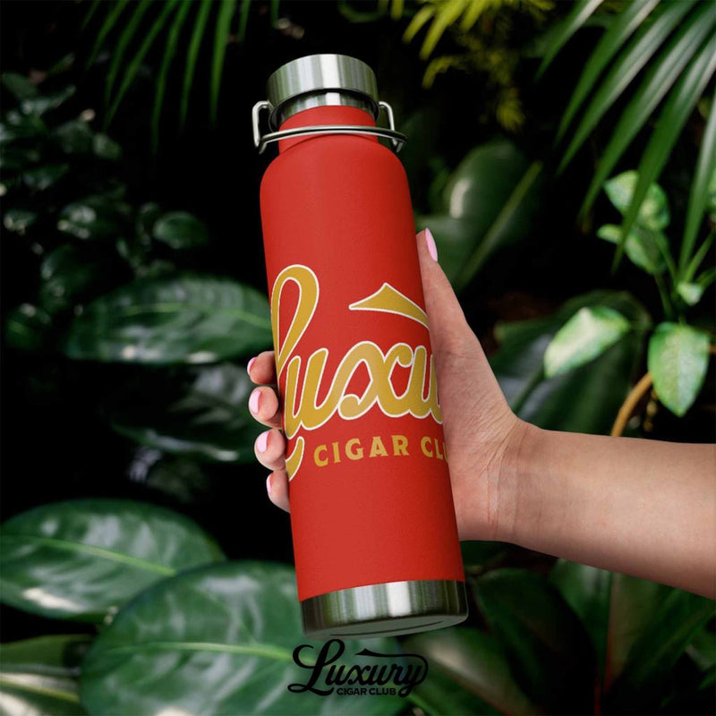 Hand holding Luxury Cigar Club copper vacuum 22oz insulated bottle in red with gold logo lifestyle view outdoors with green leaves