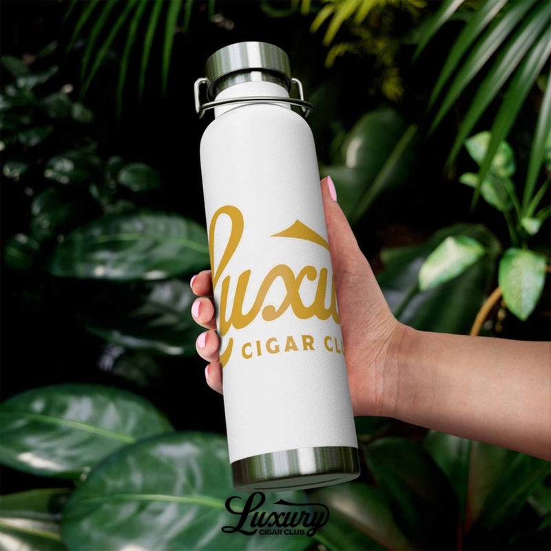 Luxury Cigar Club copper vacuum insulated bottle in white held outdoors against tropical leaves with gold logo