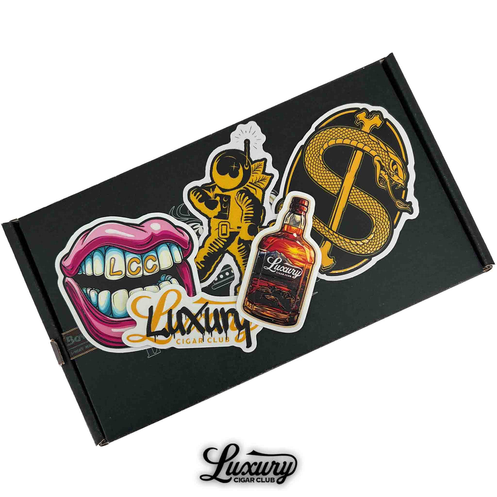 Luxury Cigar Club exclusive sticker pack featuring vibrant designs including astronaut, golden serpent dollar sign, whiskey bottle, colorful lips with “LCC,” and Luxury logo, displayed on a black background box, premium cigar accessory branding item.