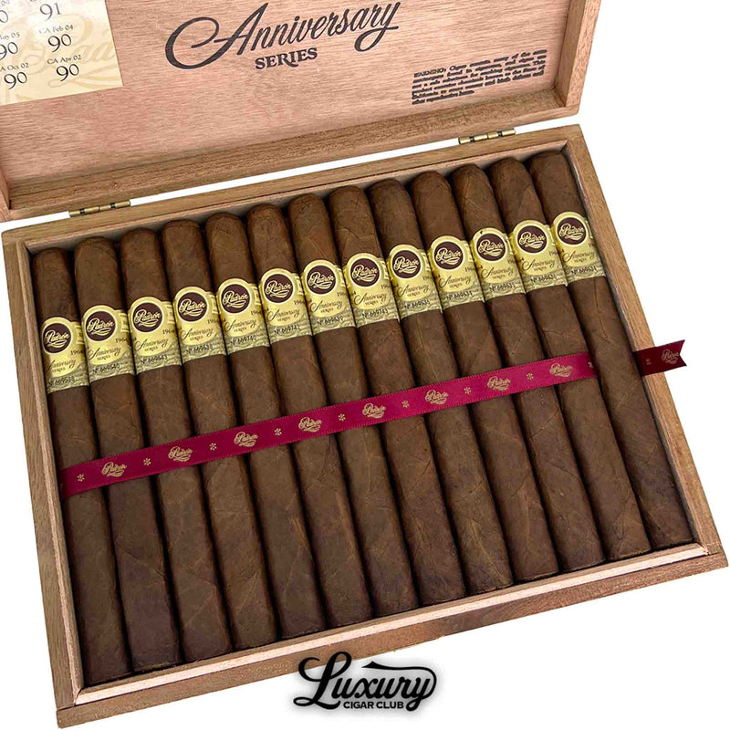 Close-up of Padron 1964 Anniversary Series Maduro Monarca cigars in their cedar box, showing dark Maduro wrappers with elegant gold embossed bands. Red ribbon detail and branded lid visible. Luxury Cigar Club logo.