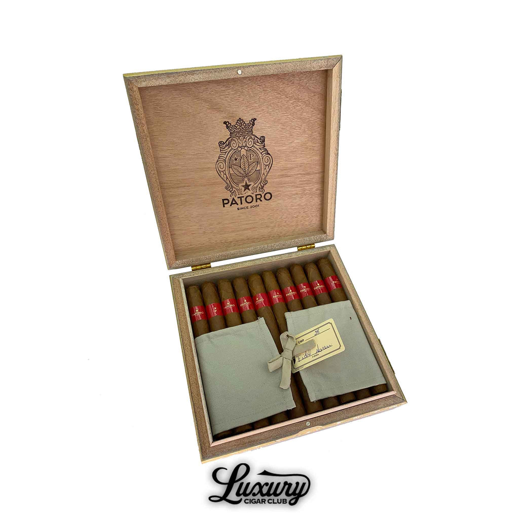 Open wooden box of Patoro Gran Añejo Reserva Churchill cigars arranged in two rows with red cigar bands and fabric pouches tied with a ribbon. The Patoro crest is engraved on the lid. Luxury Cigar Club logo displayed beneath.