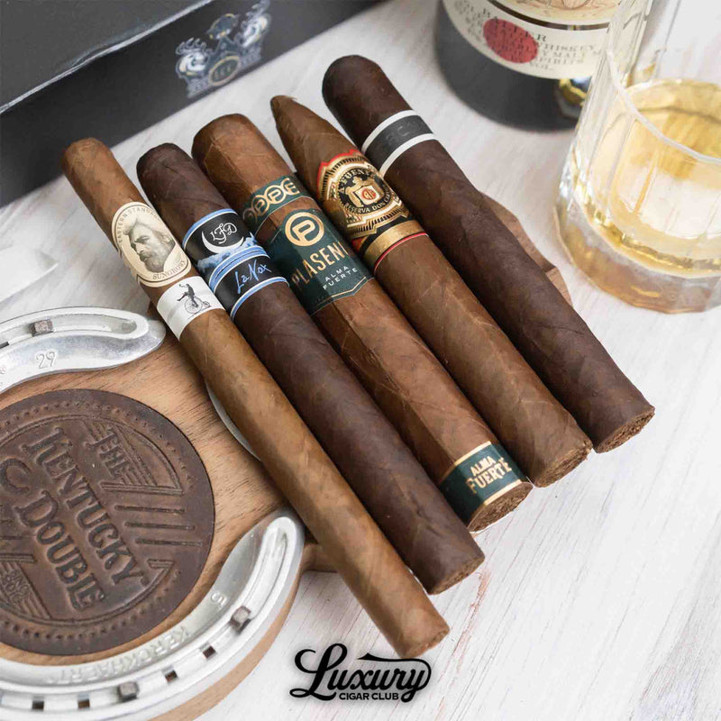 Assortment of five cigars from the Luxury Cigar Club Platinum Subscription displayed with a whiskey glass and cigar accessories, featuring Plasencia Alma Fuerte and Arturo Fuente blends.