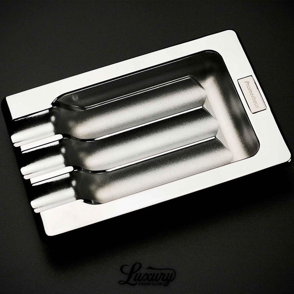 Prometheus Three Cigar Shiny Chrome Metal Ashtray featuring a sleek, polished finish with three deep cigar rests and a metallic Prometheus logo plate, displayed on a black surface above the Luxury Cigar Club logo.