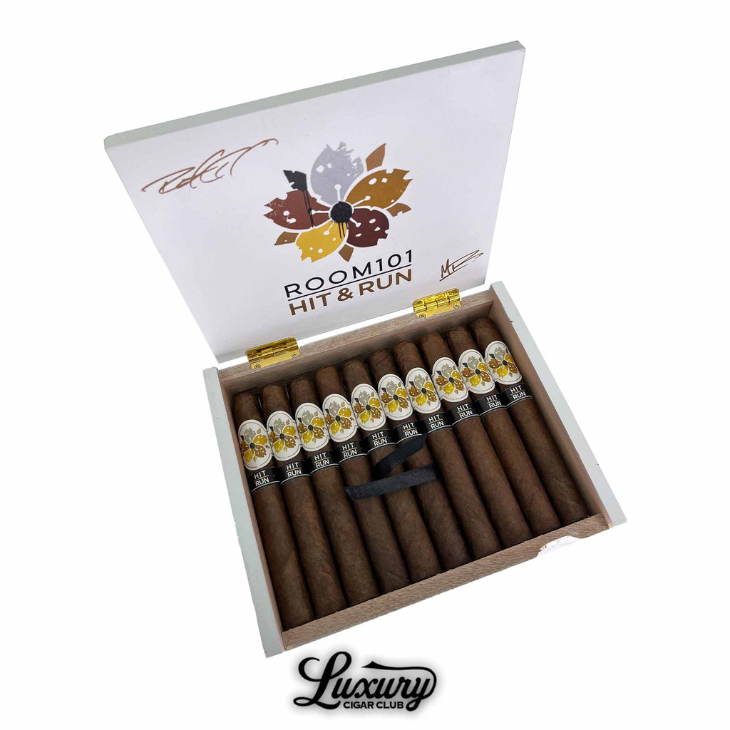 Open box of Room101 Hit and Run Toro cigars from Luxury Cigar Club showing rows of premium brown cigars with decorative white and gold floral bands arranged neatly inside
