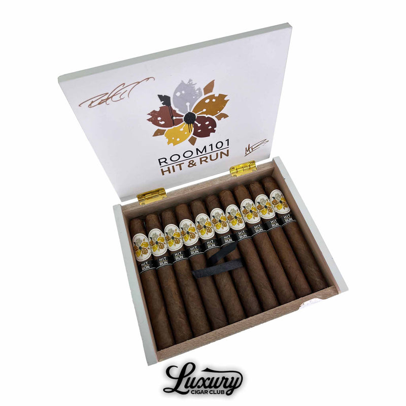 Open box of Room101 Hit and Run Toro cigars from Luxury Cigar Club showing rows of premium brown cigars with decorative white and gold floral bands arranged neatly inside