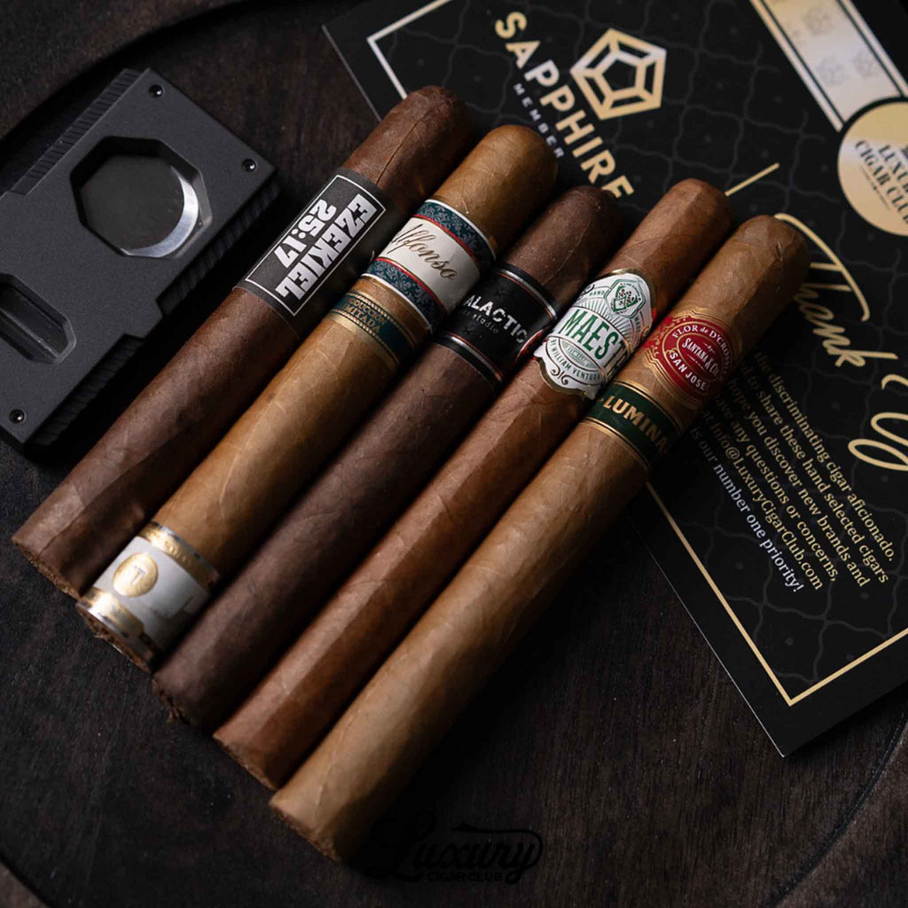 A close-up of four premium cigars from the Luxury Cigar Club Sapphire Subscription displayed on a dark surface, including recognizable labels from Alfonso, Black Works Studio, Maestros del Sabor, and Flor de D’Crossier, placed beside a cigar cutter and Sapphire membership card.