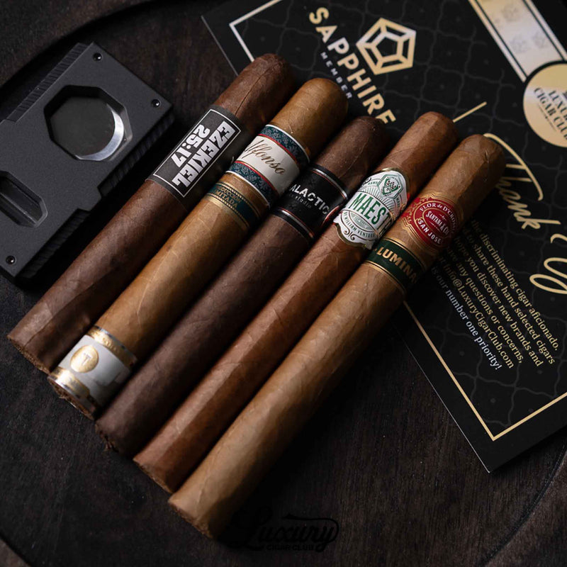 A close-up of four premium cigars from the Luxury Cigar Club Sapphire Subscription displayed on a dark surface, including recognizable labels from Alfonso, Black Works Studio, Maestros del Sabor, and Flor de D’Crossier, placed beside a cigar cutter and Sapphire membership card.