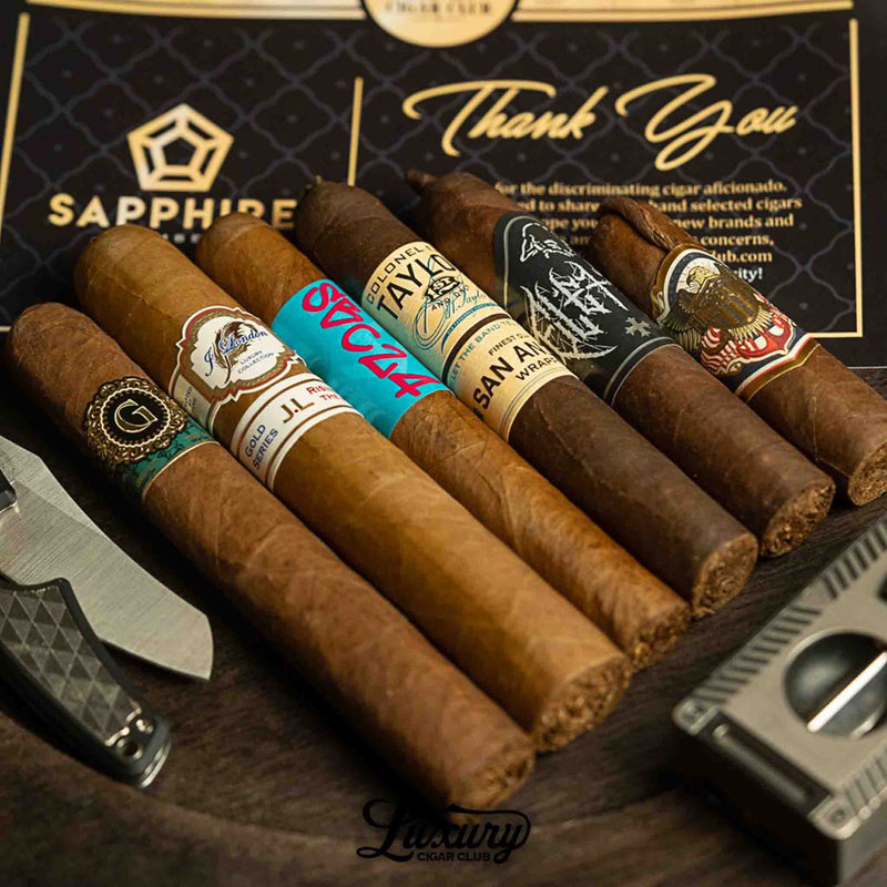 Six premium cigars from the Luxury Cigar Club Sapphire Subscription arranged neatly on a dark table, featuring vibrant cigar bands including J. London, Colonel E.H. Taylor, Freud, and other boutique labels, with a Sapphire membership thank-you card in the background.