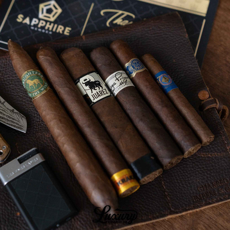 A selection of five cigars from the Luxury Cigar Club Sapphire Subscription displayed on a leather mat, featuring brands like Juarez, Fratello, and RoMa Craft, with a lighter and knife positioned nearby for an elegant presentation.