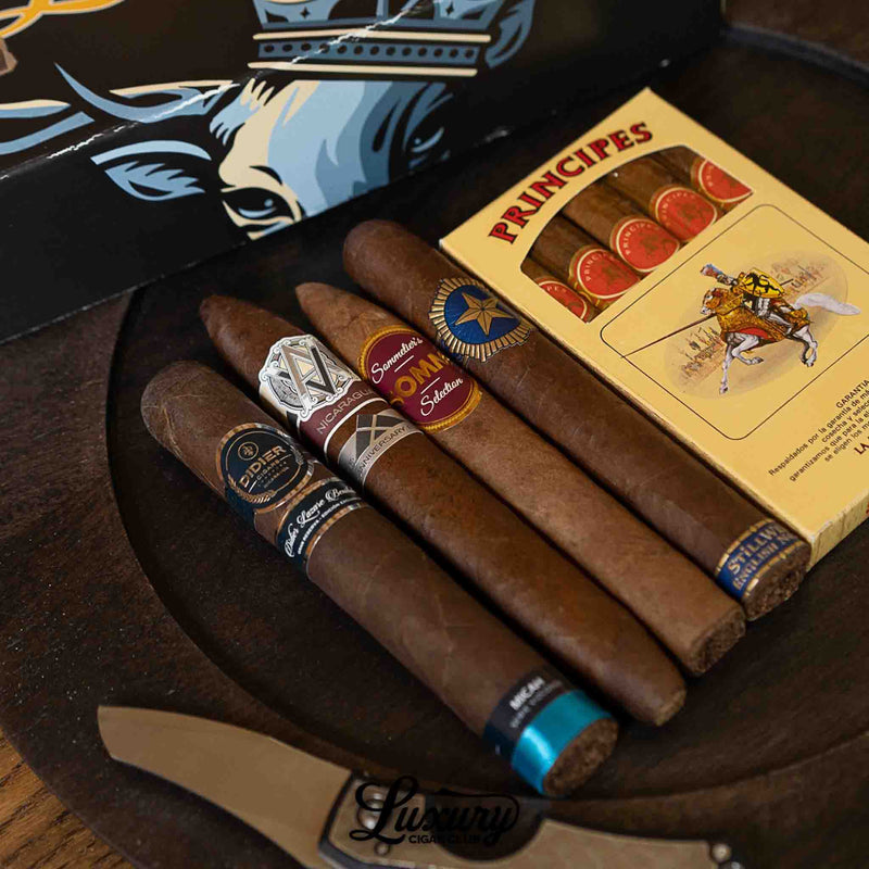 Five premium cigars from the Luxury Cigar Club Sapphire Subscription arranged beside a yellow box of Principes cigars, showcasing bands from AVO, Joya de Nicaragua, and others, with a cigar knife and decorative tray in the background.