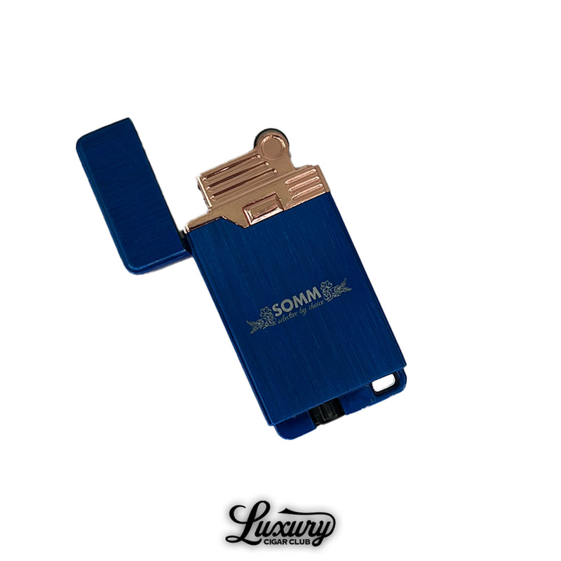 SOMM Hybrid Lighter in blue with rose gold ignition, open view showing flame mechanism, engraved SOMM logo on the body, photographed on a white background with Luxury Cigar Club logo.