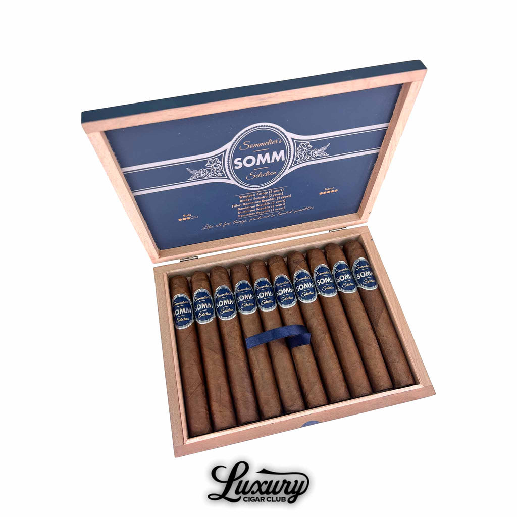 Open box of Somm Rioja Toro cigars from Luxury Cigar Club showing 12 Corojo-wrapped premium Dominican cigars with blue SOMM bands inside a wooden box.