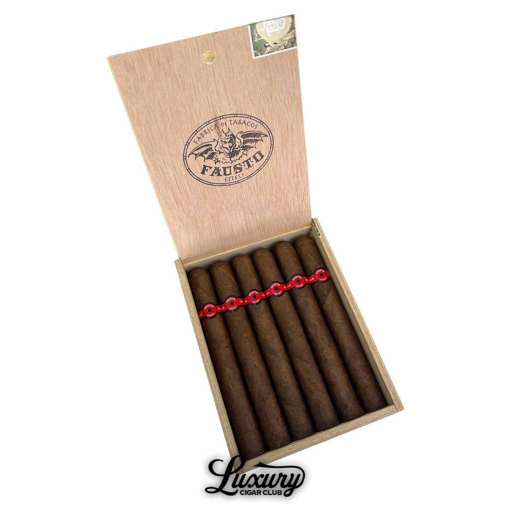 Open wooden box containing ten Tatuaje Fausto FT 187 cigars, each wrapped in a dark brown leaf and adorned with bold red and black bands. The inside lid features the "Fábrica de Tabacos Fausto Estelí" logo with a bat illustration. The Luxury Cigar Club logo is placed below the box.