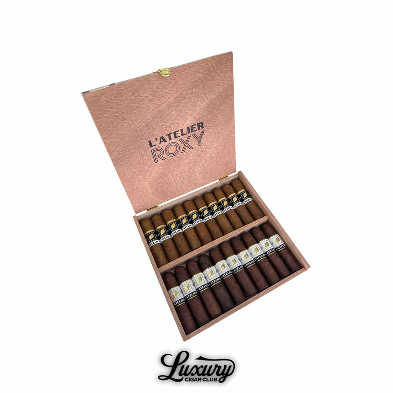 Open box of Tatuaje L’Atelier Roxy cigars showing two rows of short robusto cigars with black and white bands, presented in a wooden box on a white background with the Luxury Cigar Club logo.