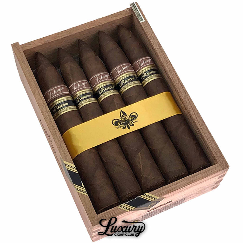 Close up photo of Tatuaje Reserva Broadleaf Unicos cigars in an open wooden box, highlighting the dark broadleaf wrappers, brown and gold Reserva bands, and the yellow fleur-de-lis ribbon holding the cigars together.