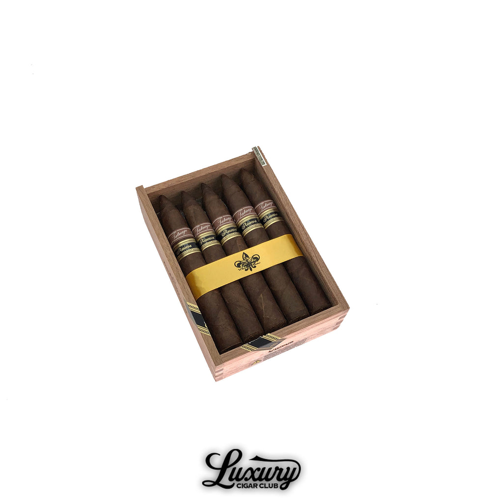 Luxury Cigar Club product photo of Tatuaje Reserva Broadleaf Unicos cigars in an open wooden box on a white background, showing six dark broadleaf torpedo cigars with brown and gold Reserva bands, secured with a yellow fleur-de-lis ribbon.
