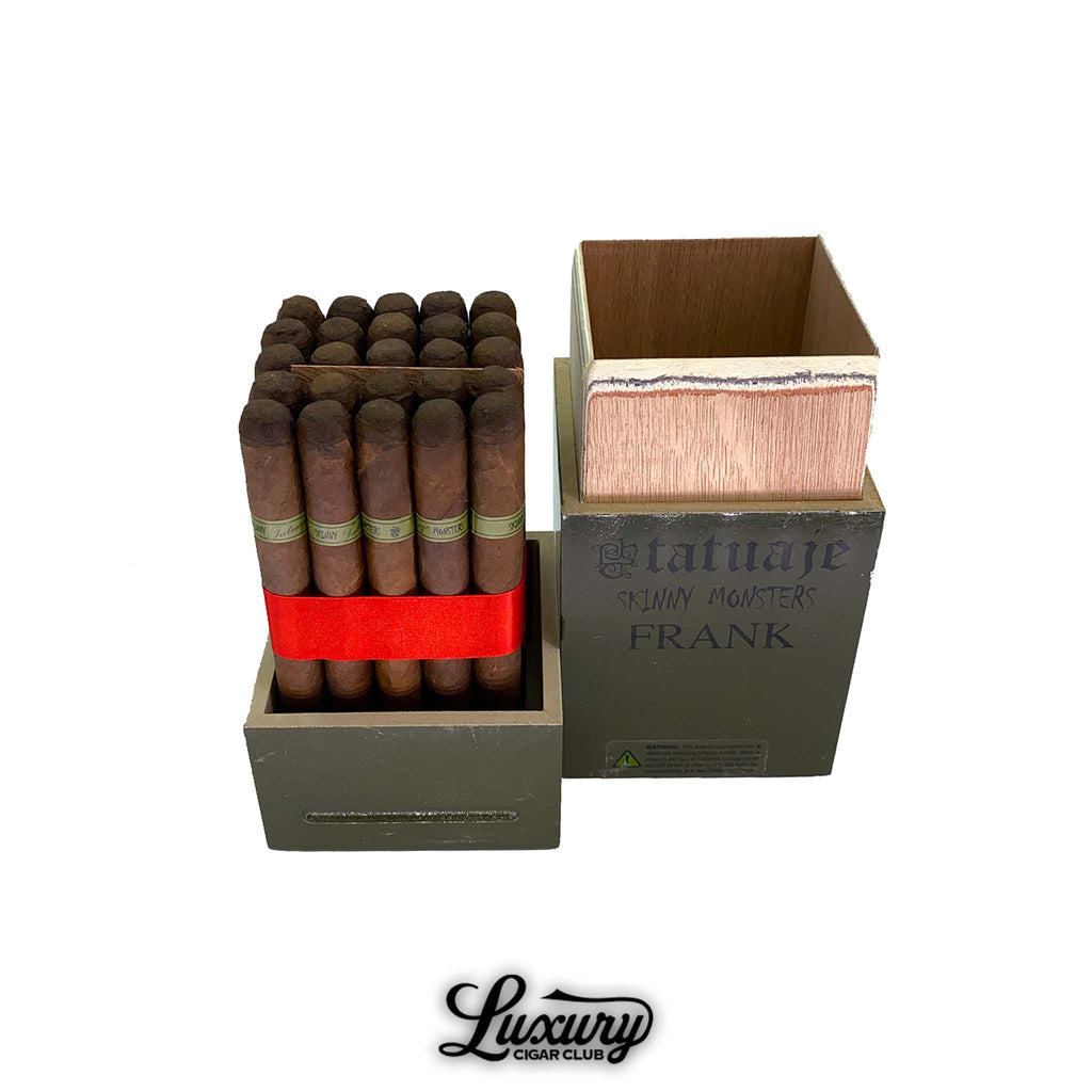 Tatuaje skinny monsters frank petit lancero cigars displayed in an open olive-green coffin-style box with the lid removed, showing a tightly packed bundle of brown cigars wrapped with a red ribbon, each cigar featuring a green tatuaje frank band, set against a white background with the luxury cigar club logo beneath