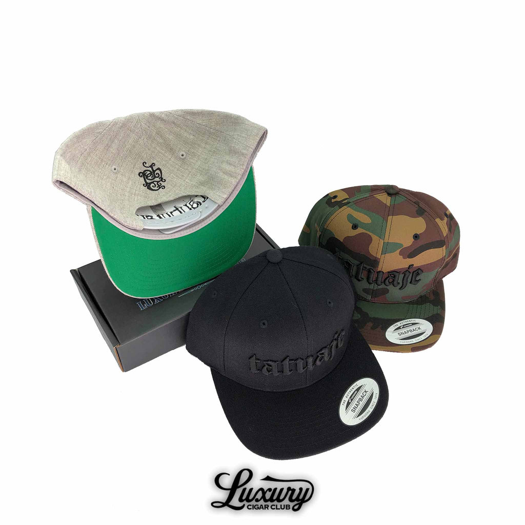 Tatuaje snapback hats in black camo and grey styles displayed with a branded box at Luxury Cigar Club.