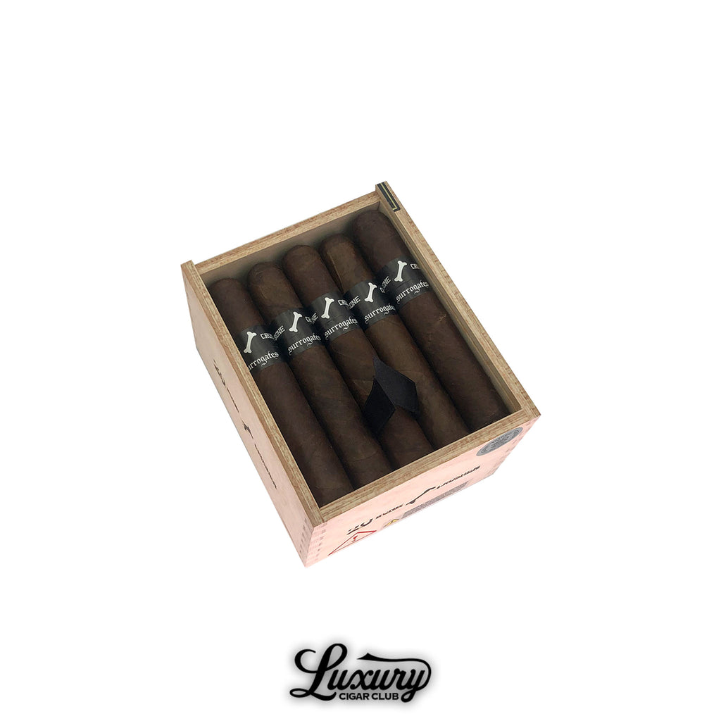 Open wooden box of Tatuaje Surrogates Bone Crusher cigars showing six dark brown robusto cigars with black bands featuring a white bone logo, displayed at an angle on a white background with Luxury Cigar Club branding.