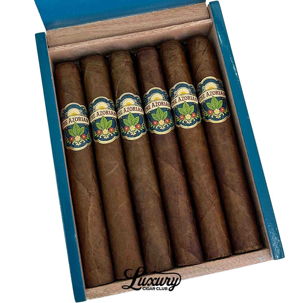 Close-up view of six Tradecraft Azorian Corona Gorda cigars resting inside an open blue presentation box, highlighting smooth brown wrappers and ornate blue, green, cream, and floral cigar bands. Luxury Cigar Club.