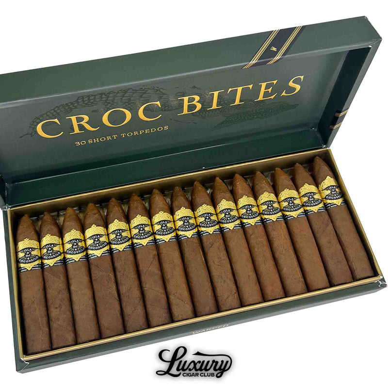 Close-up of Warped Isla del Cocodrilo Croc Bites cigars arranged in the open 30-count box, highlighting gold embossed bands and torpedo shape by Luxury Cigar Club.