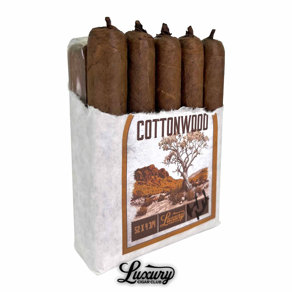 Open pack of Wildfire Cottonwood cigars showing five rustic brown cigars with pigtail caps, partially wrapped in white paper and labeled with the Cottonwood desert scene design, presented by Luxury Cigar Club.