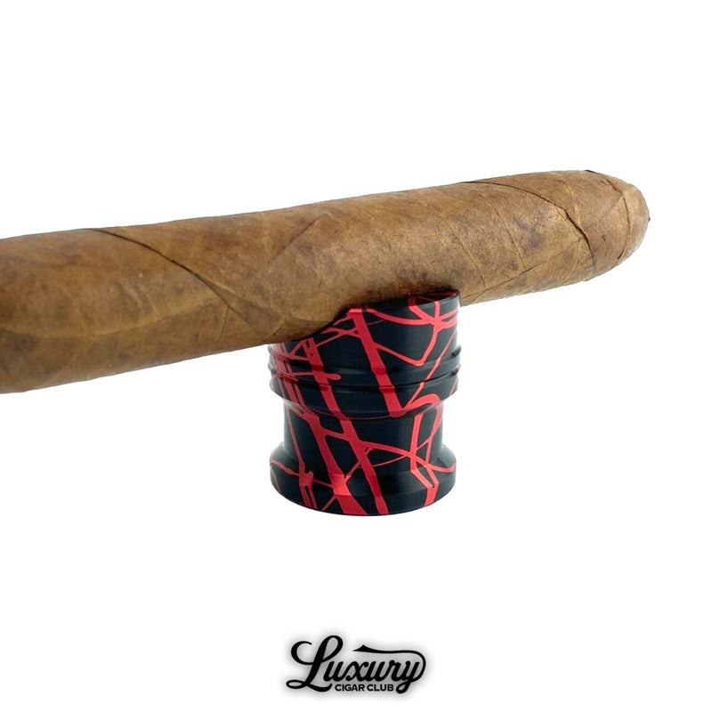 Splashed aluminum Zimmer Maverick cigar rest with red and black abstract design holding a premium cigar, photographed for Luxury Cigar Club.
